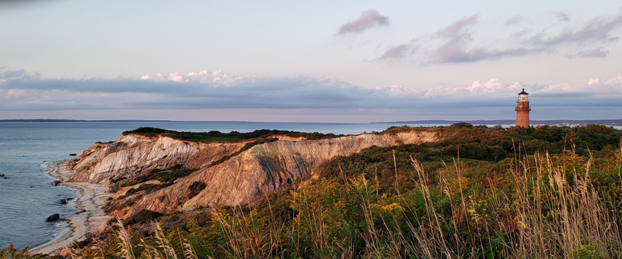 Classic lighthouse image. the sun is getting close to setting, but there is still some blue in the sky. there's a lovely hint of peach pastels on the horizon, craggy cliffs in the foreground